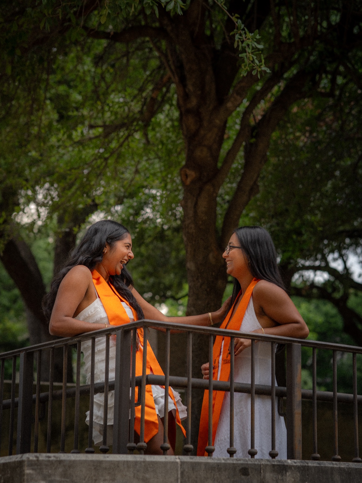 Two graduates laughing together beneath a leafy tree beside a stair railing.