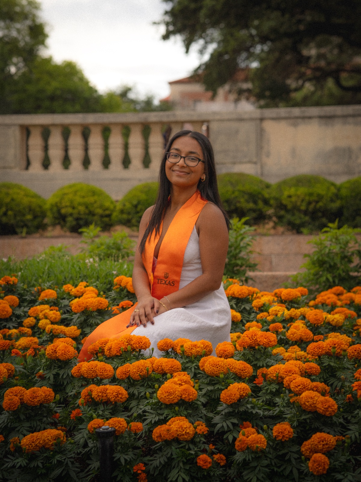 A graduate wearing glasses and an orange stole seated among orange flowers.