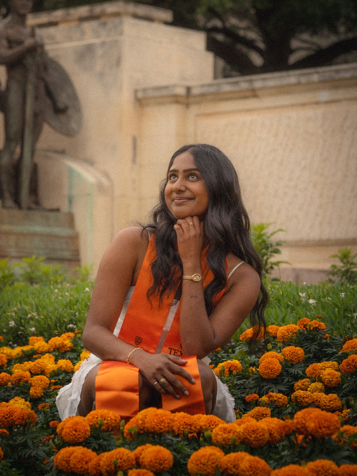 A graduate resting her chin on her hand among orange flowers by a stone wall.