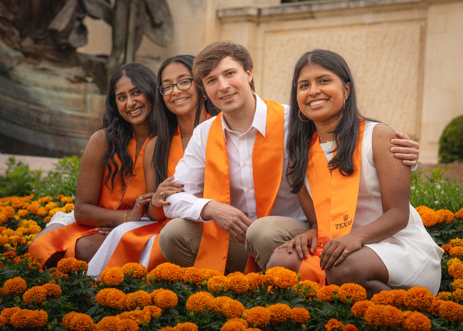 Four friends in orange graduation stoles seated together among flowers.