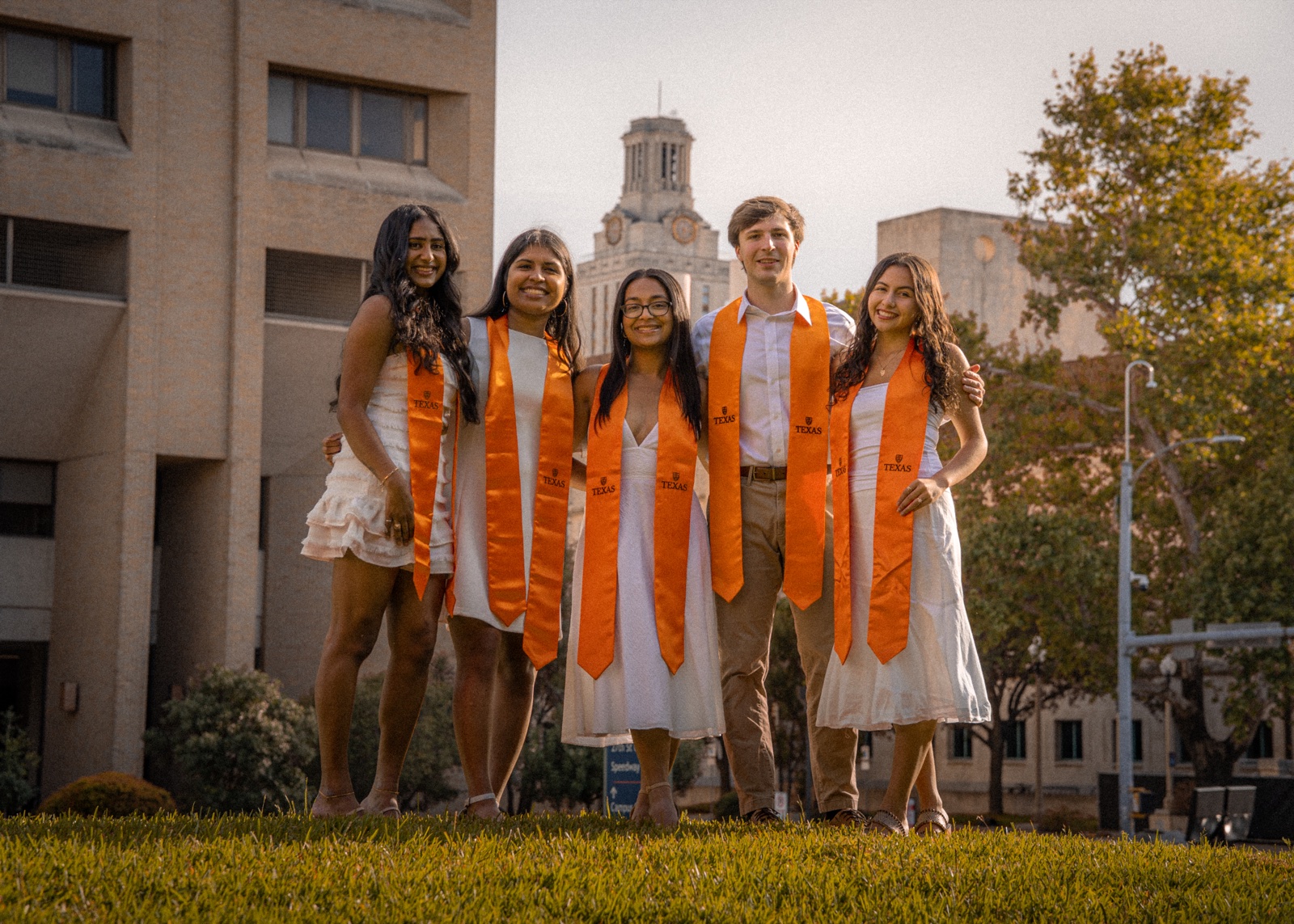 Five graduates standing together on a lawn with a campus tower in the background.