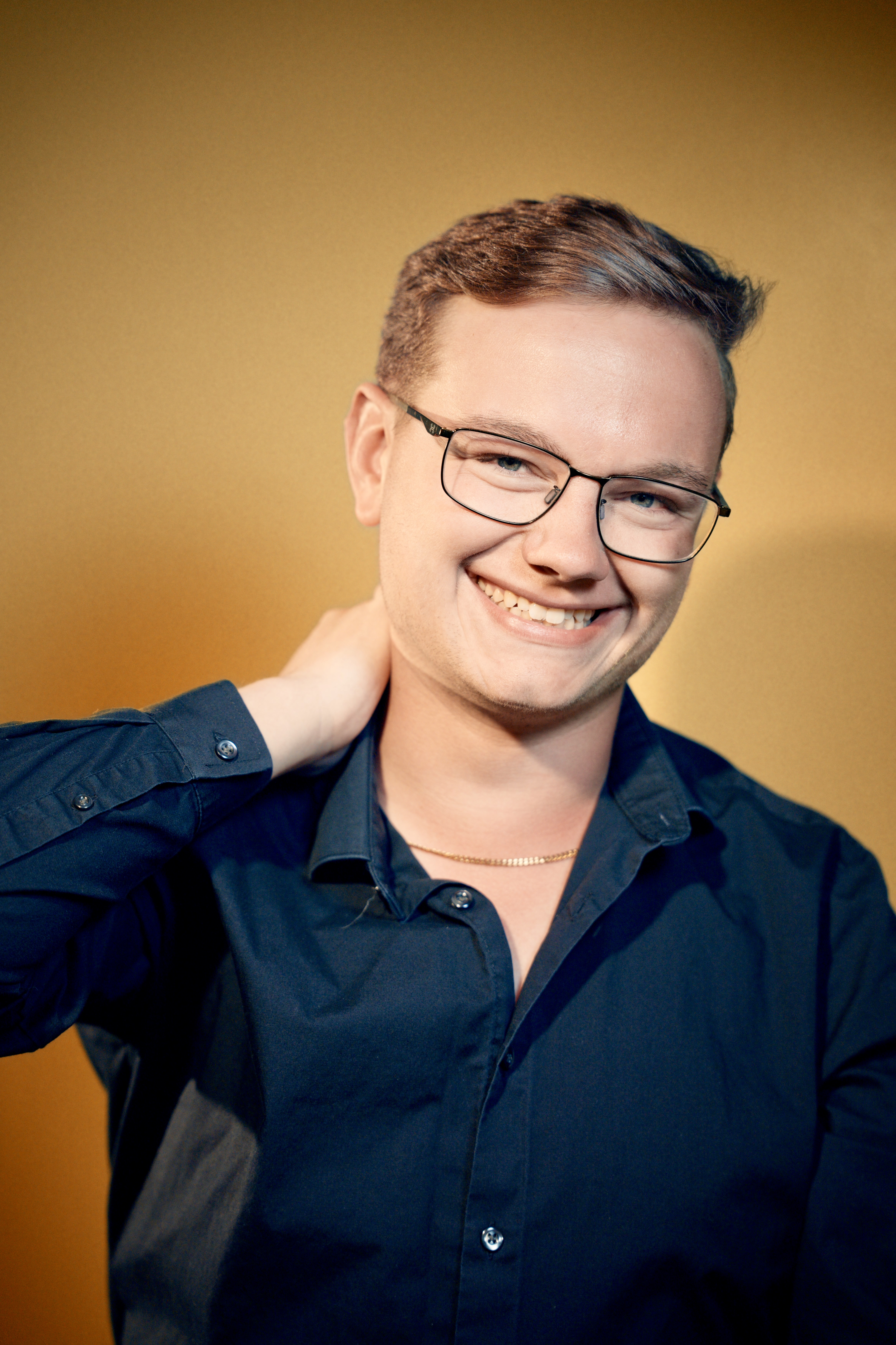 Will smiling, wearing glasses and a navy shirt against a warm gold background