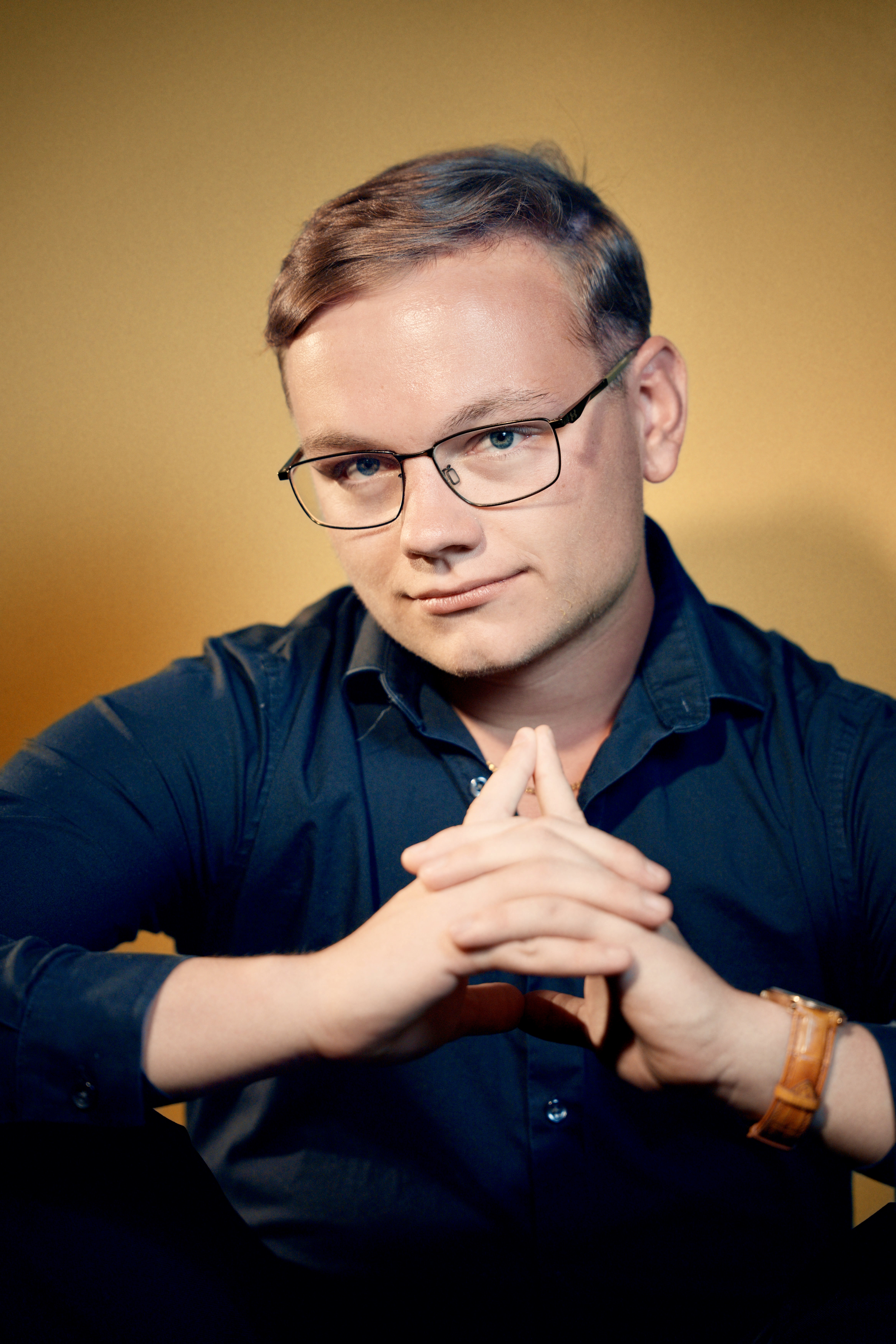 Will wearing glasses and a navy shirt, hands clasped, against a warm gold backdrop